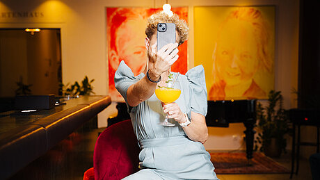 Person im hellblauen Overall macht ein Selfie an der Bar mit Cocktail in der Hand